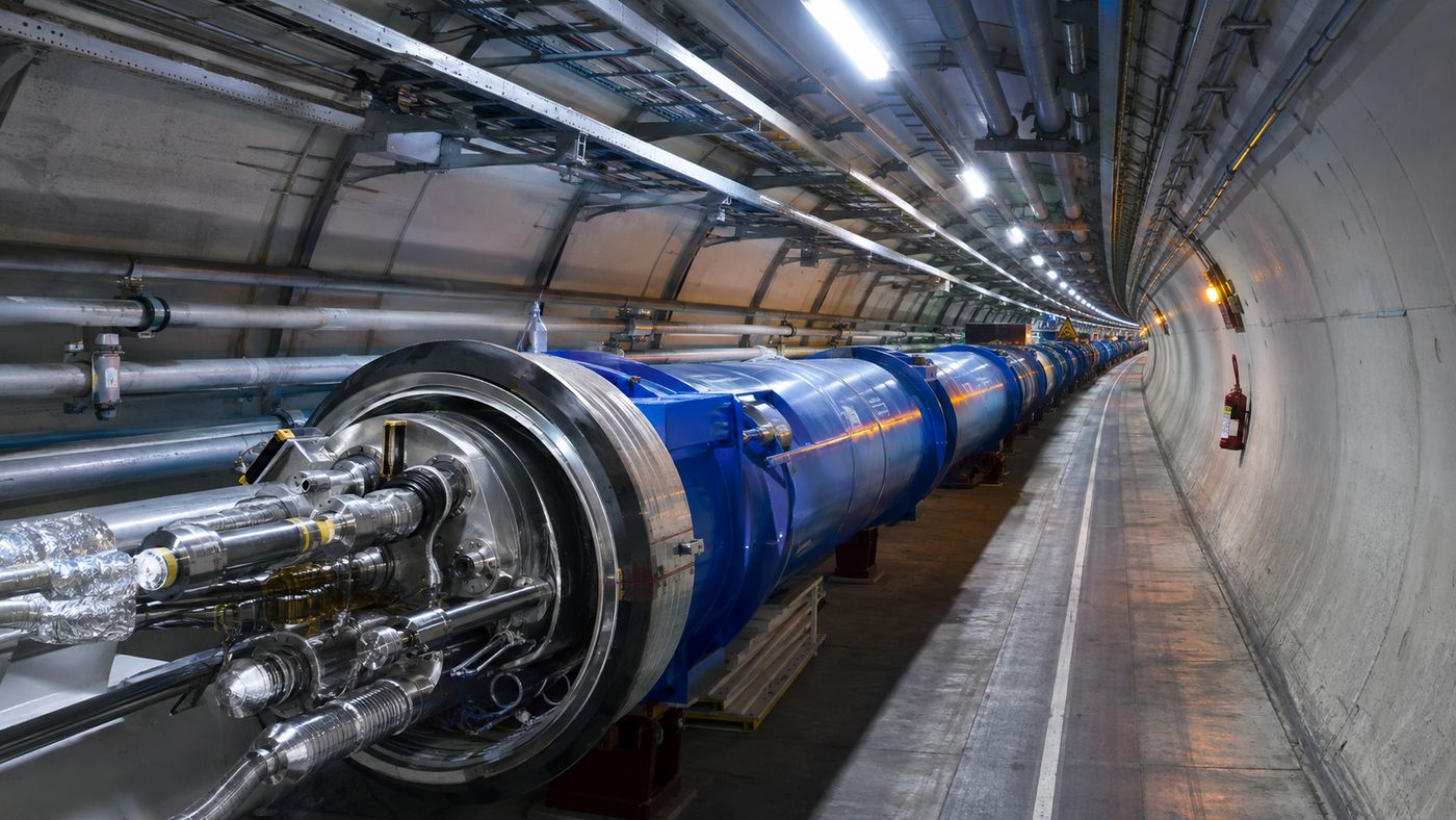 The interior of a massive underground particle accelerator tunnel, the circular tube of the beam pipe extending into the distance under bright fluorescent lighting