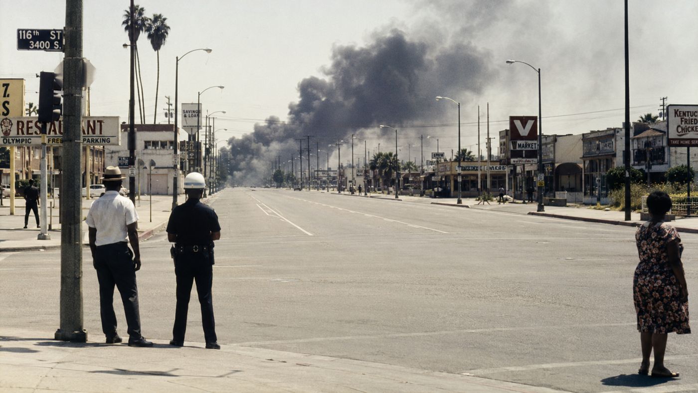 A Los Angeles urban street in the 1960s with smoke rising from buildings in the distance, the wide boulevard empty and tense in the summer heat