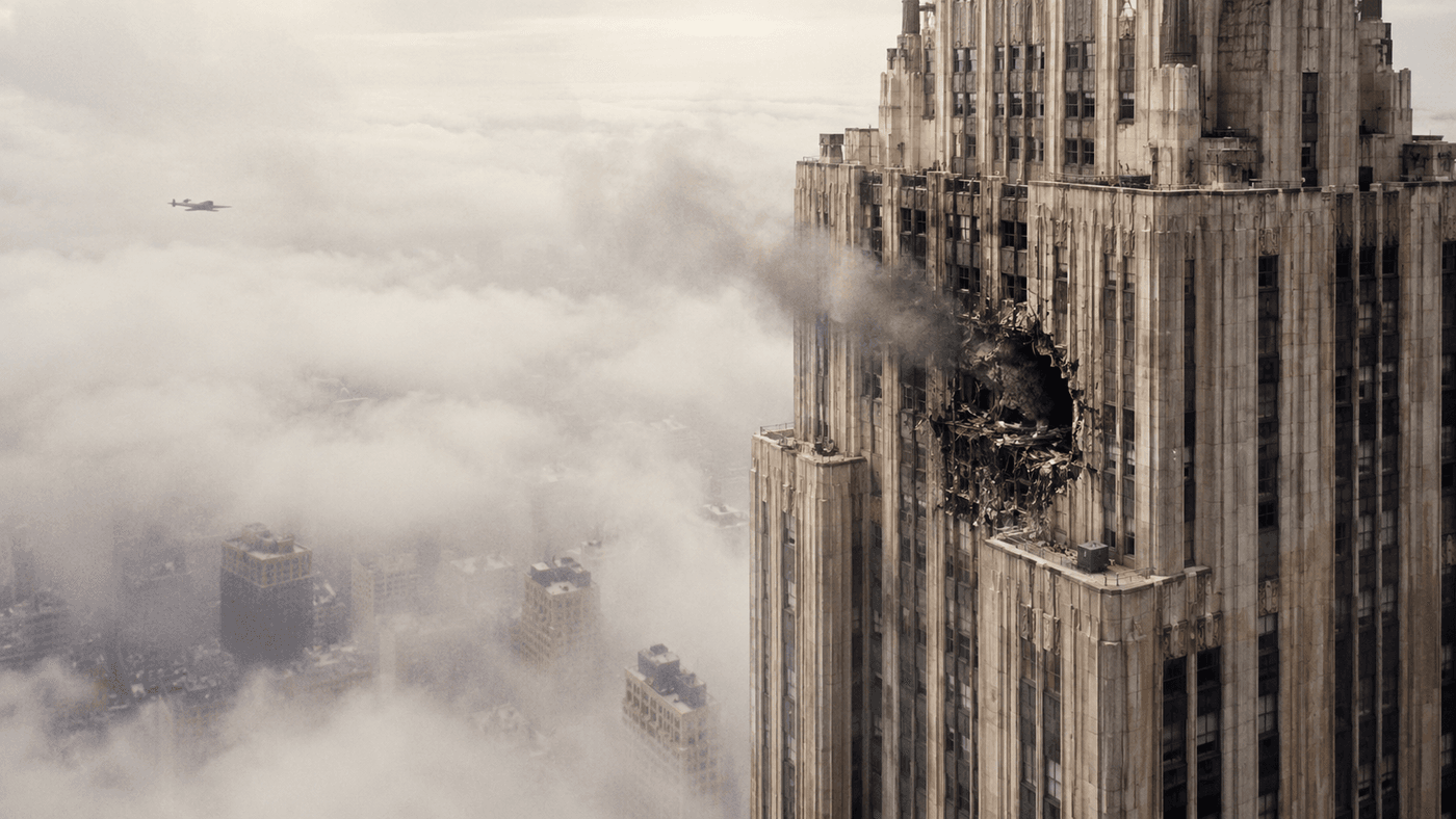 The upper floors of a towering New York skyscraper in heavy morning fog, smoke visible from a gash in the building's stone facade